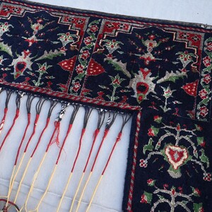 K&ouml;nnte beinhalten: Ausschnitt eines dunkelblauen Teppichs mit aufwendigen Blumenmustern in Rot, Gr&uuml;n und Wei&szlig;. Der Teppich hat einen detaillierten Rand und lange, cremefarbene Fransen mit roten Akzenten. Das Design ist symmetrisch und traditionell.