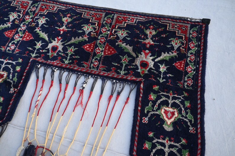 K&ouml;nnte beinhalten: Ausschnitt eines dunkelblauen Teppichs mit aufwendigen Blumenmustern in Rot, Gr&uuml;n und Wei&szlig;. Der Teppich hat einen detaillierten Rand und lange, cremefarbene Fransen mit roten Akzenten. Das Design ist symmetrisch und traditionell.