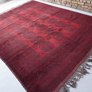 May include: A rectangular, burgundy-colored rug with a central medallion design and a patterned border. The rug features a repeating pattern of dark brown shapes and has a fringe along the edges. The rug is made of a woven material.
