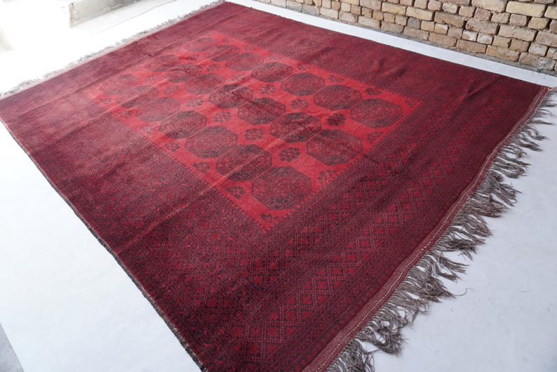 May include: A rectangular, burgundy-colored rug with a central medallion design and a patterned border. The rug features a repeating pattern of dark brown shapes and has a fringe along the edges. The rug is made of a woven material.