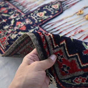 K&ouml;nnte beinhalten: Ein Abschnitt eines marineblauen Teppichs mit einem detaillierten Blumenmuster in Rot, Gr&uuml;n und Wei&szlig;. Der Rand des Teppichs ist gefaltet und zeigt einen strukturierten Rand. Der Teppich hat dekorative Quasten.