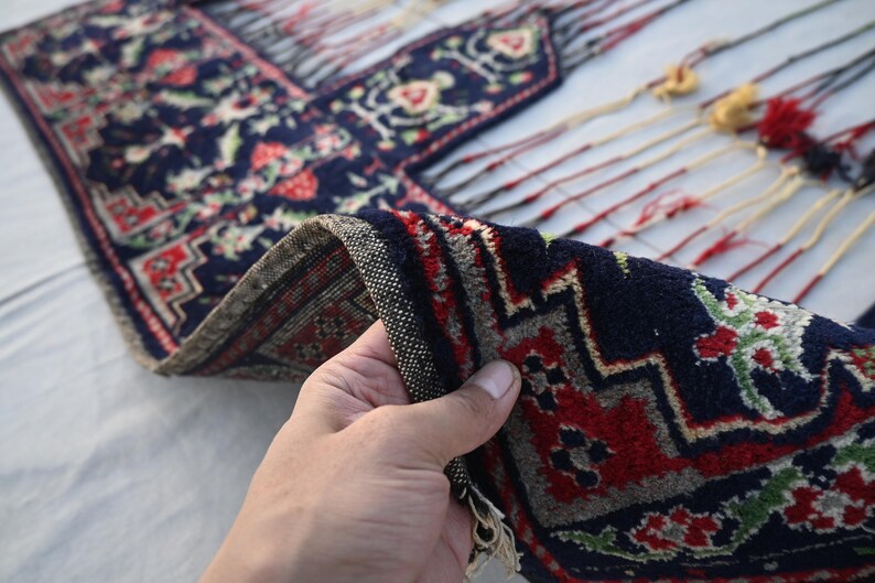 K&ouml;nnte beinhalten: Ein Abschnitt eines marineblauen Teppichs mit einem detaillierten Blumenmuster in Rot, Gr&uuml;n und Wei&szlig;. Der Rand des Teppichs ist gefaltet und zeigt einen strukturierten Rand. Der Teppich hat dekorative Quasten.