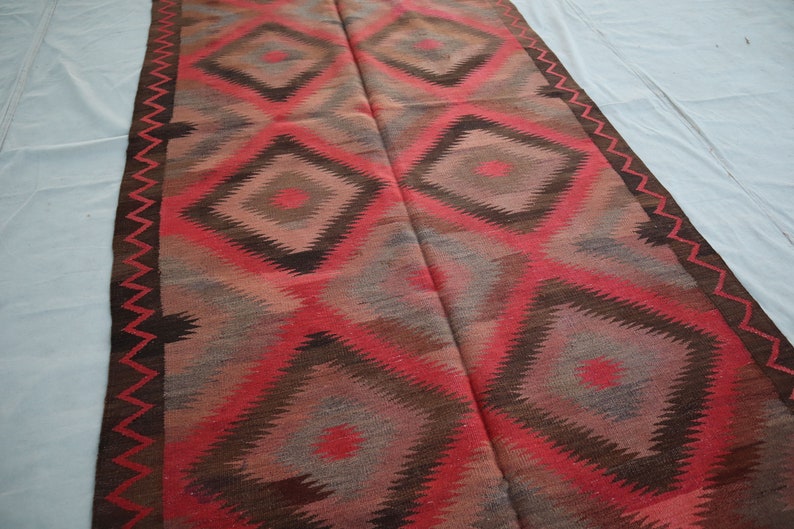 May include: A red, brown, and gray patterned rug with a geometric diamond design. The rug has a fringed edge.