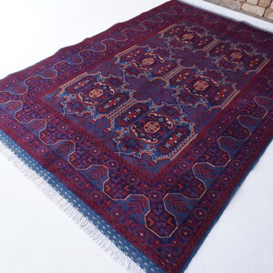 May include: A rectangular, hand-knotted rug with a deep purple field and intricate geometric patterns in red, blue, and cream. The rug features a detailed border and fringed edges, showcasing traditional craftsmanship.