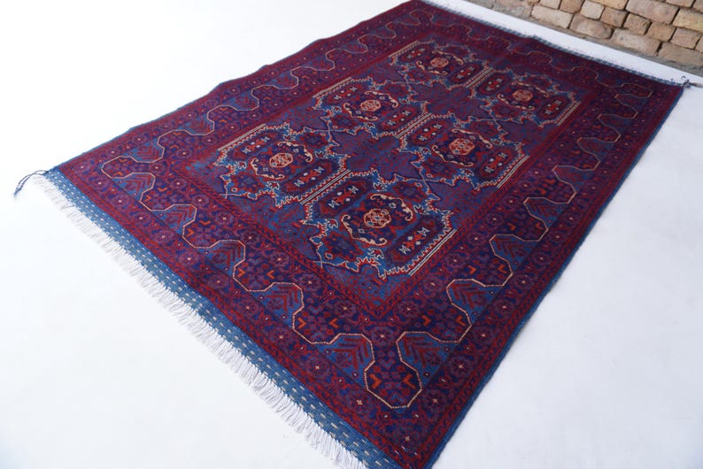 May include: A rectangular, hand-knotted rug with a deep purple field and intricate geometric patterns in red, blue, and cream. The rug features a detailed border and fringed edges, showcasing traditional craftsmanship.