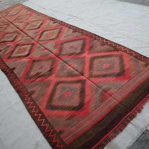 May include: A long, rectangular rug with a geometric pattern of diamonds in shades of red, brown, and gray. The rug has a fringe border.
