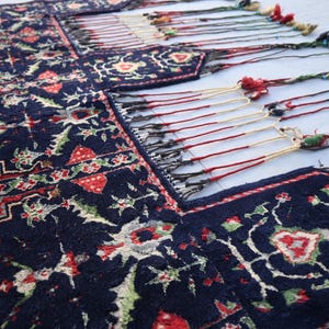 Puede incluir: Primer plano de una alfombra azul oscuro con un dise&ntilde;o floral en rojo, verde y blanco. La alfombra tiene un borde con flecos y borlas de colores. El patr&oacute;n es detallado, lo que sugiere una construcci&oacute;n tejida a mano.