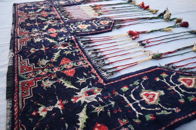 Puede incluir: Primer plano de una alfombra azul oscuro con un dise&ntilde;o floral en rojo, verde y blanco. La alfombra tiene un borde con flecos y borlas de colores. El patr&oacute;n es detallado, lo que sugiere una construcci&oacute;n tejida a mano.