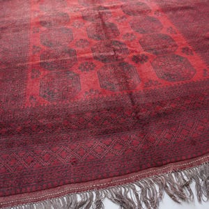 May include: A rectangular, hand-woven rug with a deep red field and a darker red border. The central design features repeating geometric patterns in black. The rug has a fringe along one edge, adding texture and detail. This rug is suitable for home decor.