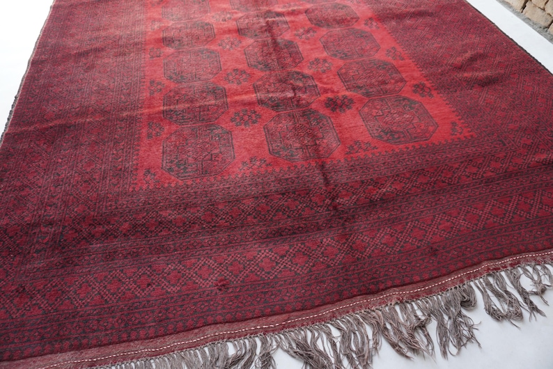 May include: A rectangular, hand-woven rug with a deep red field and a darker red border. The central design features repeating geometric patterns in black. The rug has a fringe along one edge, adding texture and detail. This rug is suitable for home decor.