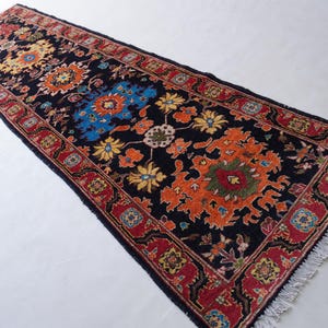 May include: A rectangular, patterned rug with a black background. The rug features floral designs in orange, yellow, and blue, framed by a red border. The rug has a fringed edge.