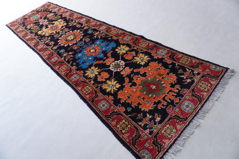 May include: A rectangular, patterned rug with a black background. The rug features floral designs in orange, yellow, and blue, framed by a red border. The rug has a fringed edge.