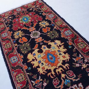 May include: A rectangular, dark blue rug with a detailed floral pattern. The rug features a central design in yellow, orange, and blue, surrounded by red borders with smaller floral motifs. The rug has a fringed edge.