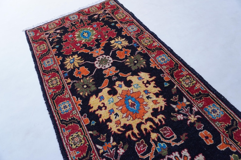 May include: A rectangular, dark blue rug with a detailed floral pattern. The rug features a central design in yellow, orange, and blue, surrounded by red borders with smaller floral motifs. The rug has a fringed edge.
