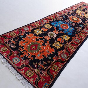 May include: A rectangular, dark blue runner rug with a detailed floral pattern. The rug features a central design in orange, yellow, and blue, framed by a red border with intricate patterns. The rug has a fringe on both ends.