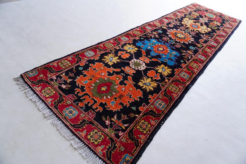 May include: A rectangular, dark blue runner rug with a detailed floral pattern. The rug features a central design in orange, yellow, and blue, framed by a red border with intricate patterns. The rug has a fringe on both ends.