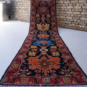 May include: A long, rectangular rug with a black background and intricate floral patterns in red, orange, yellow, and blue. The rug has a red border and white fringe.