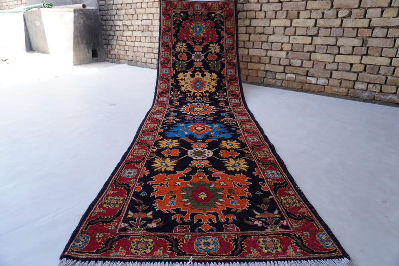 May include: A long, rectangular rug with a black background and intricate floral patterns in red, orange, yellow, and blue. The rug has a red border and white fringe.