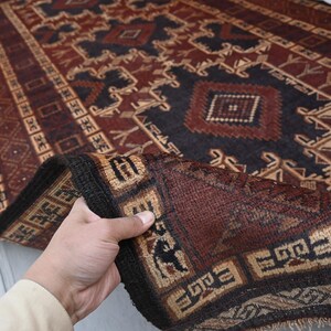 May include: A hand-woven rug with a geometric pattern in shades of burgundy, navy, and tan. The rug features a central design with diamond and rectangular shapes, and a border with repeating motifs. The rug is folded over, revealing the intricate details of the weave.