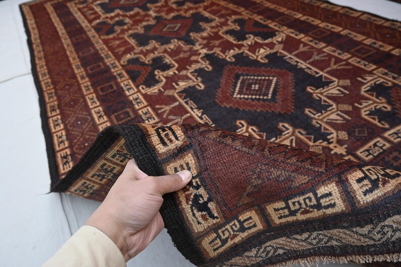 May include: A hand-woven rug with a geometric pattern in shades of burgundy, navy, and tan. The rug features a central design with diamond and rectangular shapes, and a border with repeating motifs. The rug is folded over, revealing the intricate details of the weave.