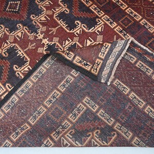 May include: A detailed shot of a patterned rug with a folded corner. The rug features a complex design with geometric shapes and repeating patterns in shades of burgundy, navy blue, and beige. The edges are finished with a dark border and fringe.