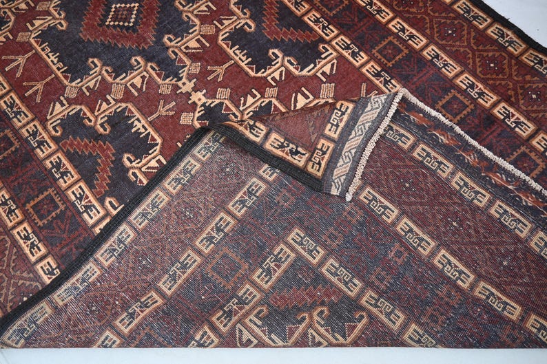 May include: A detailed shot of a patterned rug with a folded corner. The rug features a complex design with geometric shapes and repeating patterns in shades of burgundy, navy blue, and beige. The edges are finished with a dark border and fringe.