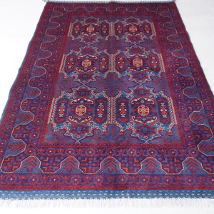 May include: A rectangular rug with a detailed, symmetrical pattern in shades of burgundy, blue, and cream. The rug features a central design with repeating floral and geometric motifs, framed by a border with a similar pattern. The rug has a fringe on one end.