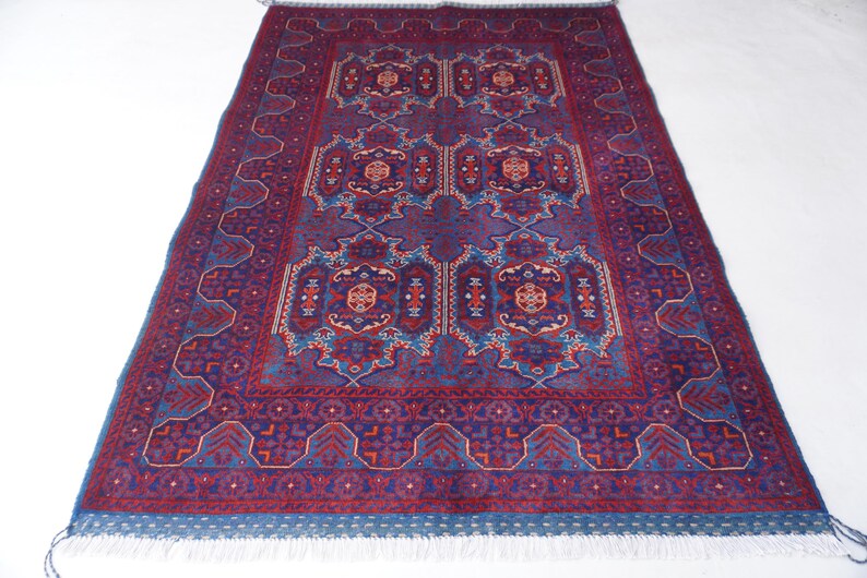 May include: A rectangular rug with a detailed, symmetrical pattern in shades of burgundy, blue, and cream. The rug features a central design with repeating floral and geometric motifs, framed by a border with a similar pattern. The rug has a fringe on one end.