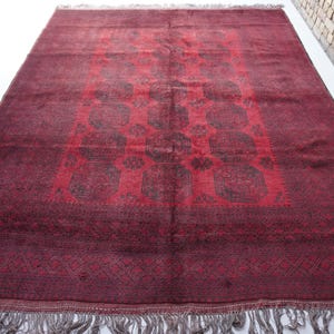 May include: A rectangular, hand-woven rug with a deep red field and intricate geometric patterns in darker shades. The rug features a central design with repeating motifs and a decorative border. The edges are finished with a fringe.