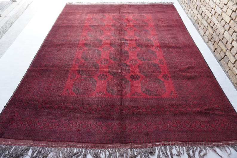 May include: A rectangular, hand-woven rug with a deep red field and intricate geometric patterns in darker shades. The rug features a central design with repeating motifs and a decorative border. The edges are finished with a fringe.