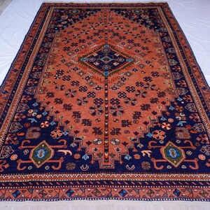 May include: A large, rectangular rug with an intricate pattern. The rug features a central field of reddish-orange with a navy blue border. The design includes floral and geometric motifs in various colors, creating a detailed and ornate appearance.