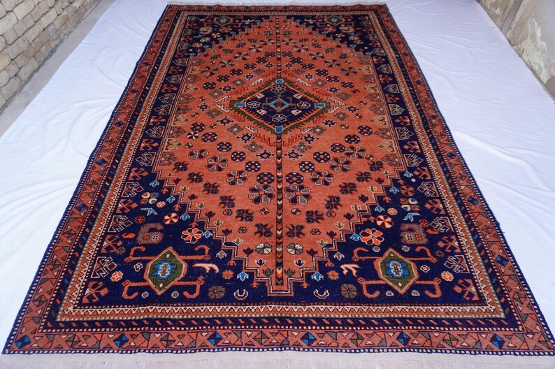 May include: A large, rectangular rug with an intricate pattern. The rug features a central field of reddish-orange with a navy blue border. The design includes floral and geometric motifs in various colors, creating a detailed and ornate appearance.