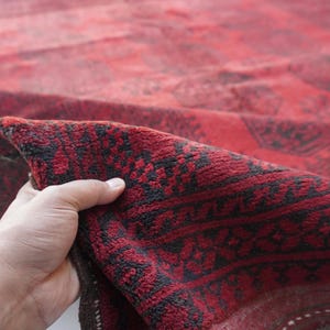 May include: A section of a red and black patterned rug is held up, revealing the intricate design. The rug's color palette is dominated by deep reds and blacks, with a detailed, geometric pattern.