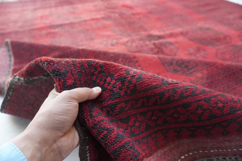 May include: A section of a red and black patterned rug is held up, revealing the intricate design. The rug's color palette is dominated by deep reds and blacks, with a detailed, geometric pattern.