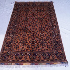 May include: Rectangular rug with a central field of rust-orange, featuring a repeating geometric pattern in navy blue and brown. The border is a darker navy with intricate designs. The rug has a fringed edge.