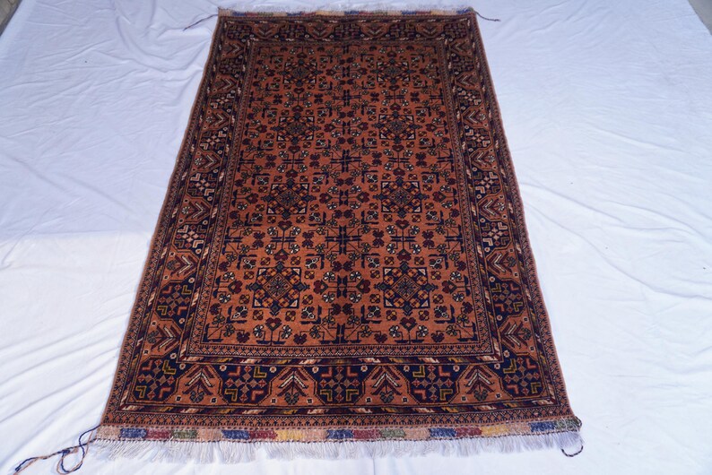 May include: Rectangular rug with a central field of rust-orange, featuring a repeating geometric pattern in navy blue and brown. The border is a darker navy with intricate designs. The rug has a fringed edge.