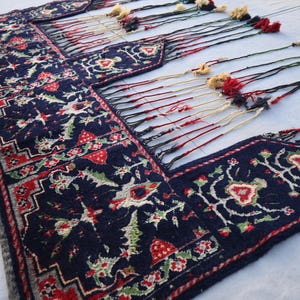 K&ouml;nnte beinhalten: Ein marineblauer, rechteckiger Teppich mit einem detaillierten Blumenmuster in Rot, Gr&uuml;n und Wei&szlig;. Der Teppich hat einen dekorativen Rand und lange Quasten in verschiedenen Farben. Der Teppich ist wahrscheinlich handgefertigt und f&uuml;r die Inneneinrichtung geeignet.