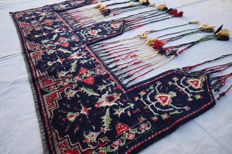 K&ouml;nnte beinhalten: Ein marineblauer, rechteckiger Teppich mit einem detaillierten Blumenmuster in Rot, Gr&uuml;n und Wei&szlig;. Der Teppich hat einen dekorativen Rand und lange Quasten in verschiedenen Farben. Der Teppich ist wahrscheinlich handgefertigt und f&uuml;r die Inneneinrichtung geeignet.