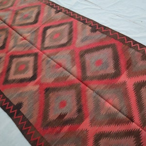 May include: A long, narrow rug with a geometric pattern in shades of red, brown, and gray. The rug has a diamond pattern with a zig-zag border.
