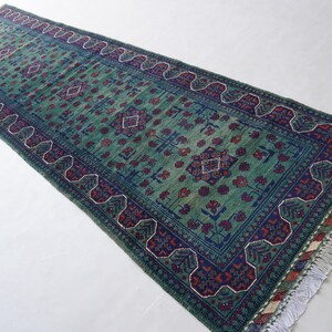 May include: A rectangular, green runner rug with a detailed pattern. The rug features a central field of green with a repeating floral design in red and blue. The border is a darker blue with a repeating geometric pattern. The rug has a fringe on the ends.