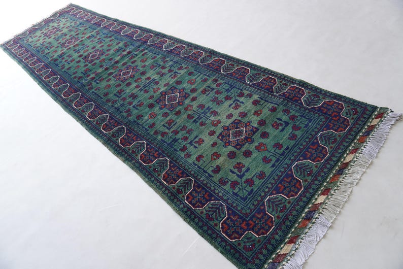 May include: A rectangular, green runner rug with a detailed pattern. The rug features a central field of green with a repeating floral design in red and blue. The border is a darker blue with a repeating geometric pattern. The rug has a fringe on the ends.