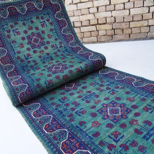 May include: A rolled-up, rectangular, teal-colored rug with a detailed geometric pattern in navy blue and red. The rug has a border with a repeating design and is displayed on a white surface.