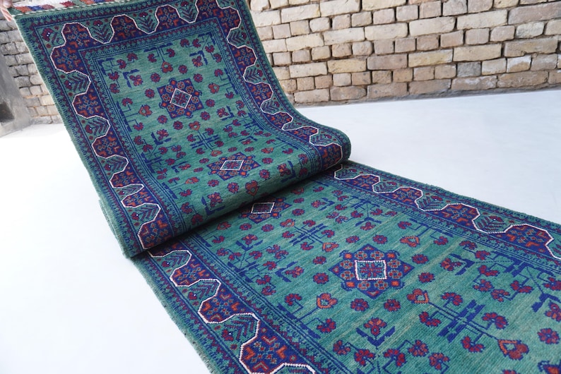 May include: A rolled-up, rectangular, teal-colored rug with a detailed geometric pattern in navy blue and red. The rug has a border with a repeating design and is displayed on a white surface.