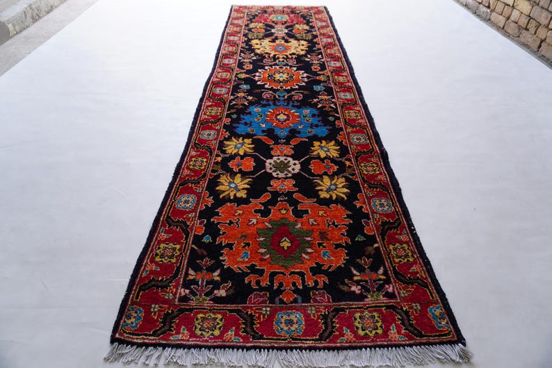 May include: A long, rectangular rug with a black background and intricate floral patterns in red, orange, yellow, and blue. The rug has a red border and white fringe on the ends. This decorative rug is suitable for a hallway or entryway.