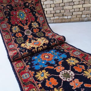 May include: A rolled-up, rectangular rug with a dark blue background and a floral pattern in red, orange, yellow, and blue. The rug has a red border with intricate designs. The rug is on a white surface with a brick wall in the background.