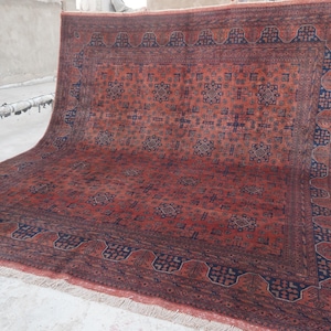 May include: A large rectangular area rug featuring a deep red base color with intricate geometric patterns in dark blue. The rug has a wide decorative border with repeating motifs and a fringe along the bottom edge, displaying a dense woven texture.