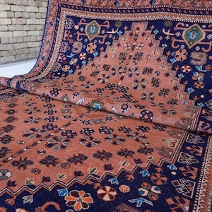 May include: An intricately patterned rug with a rich color palette of navy blue and rust orange. The rug features a repeating floral and geometric design, with a detailed border. The rug is partially rolled, showcasing its texture and design.
