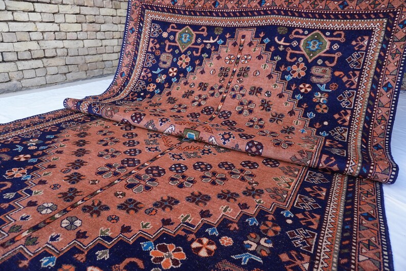 May include: An intricately patterned rug with a rich color palette of navy blue and rust orange. The rug features a repeating floral and geometric design, with a detailed border. The rug is partially rolled, showcasing its texture and design.