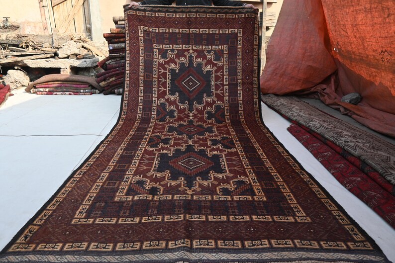May include: A rectangular, patterned rug with a central geometric design in shades of brown, black, and beige. The rug features intricate borders and a symmetrical pattern, suitable for home decor. The rug is displayed outdoors.