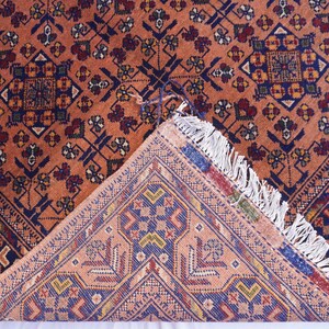May include: An intricately patterned rug with a warm, reddish-orange background. The design features geometric shapes in navy blue, burgundy, and cream. The rug's corner is folded back, revealing a detailed underside with a similar color palette and fringe.